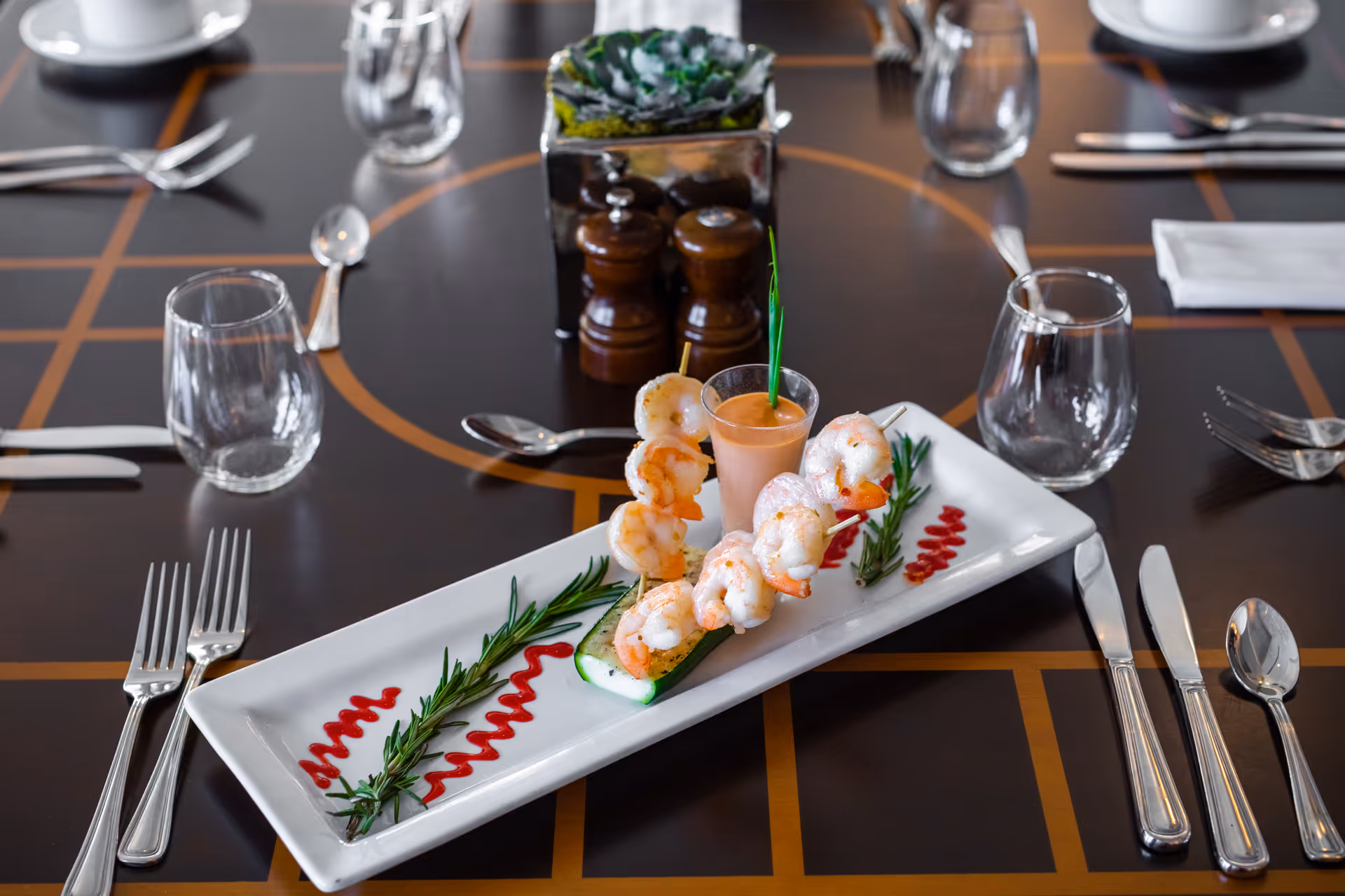 A rectangular white plate with shrimp skewers arranged on a cucumber slice, garnished with rosemary sprigs and red sauce, accompanied by a small glass of dipping sauce, placed on a dark wooden dining table set with silverware, glasses, and napkins.