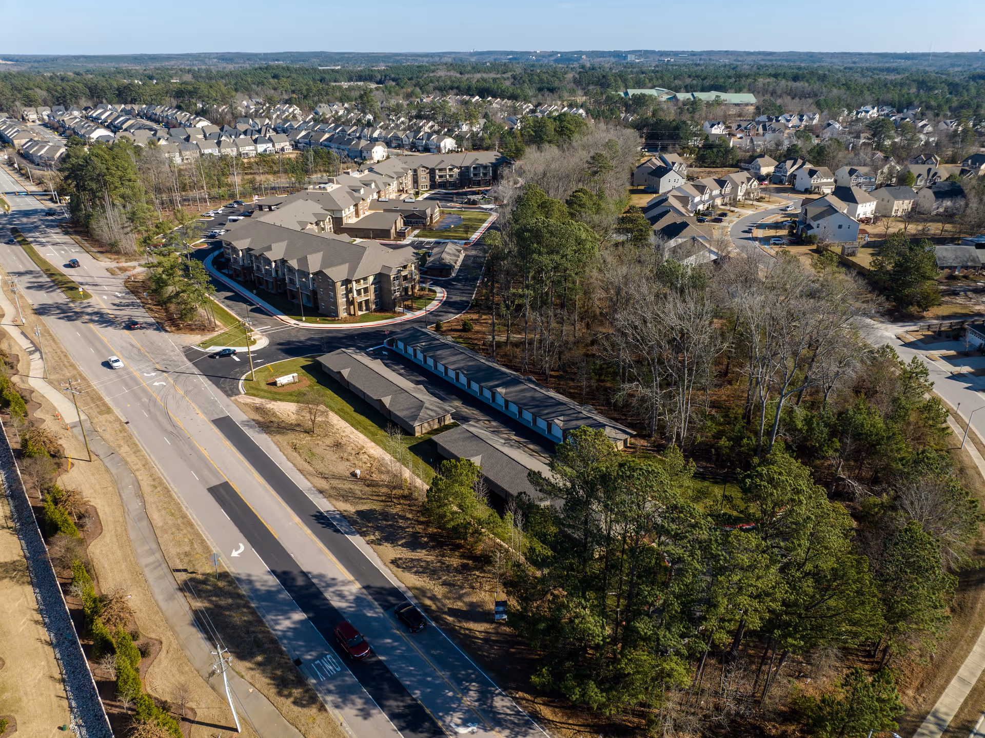 Aerial view of a residential area featuring multiple apartment buildings, houses, and a main road with cars. The scene includes trees and wooded areas surrounding the buildings under a clear sky.