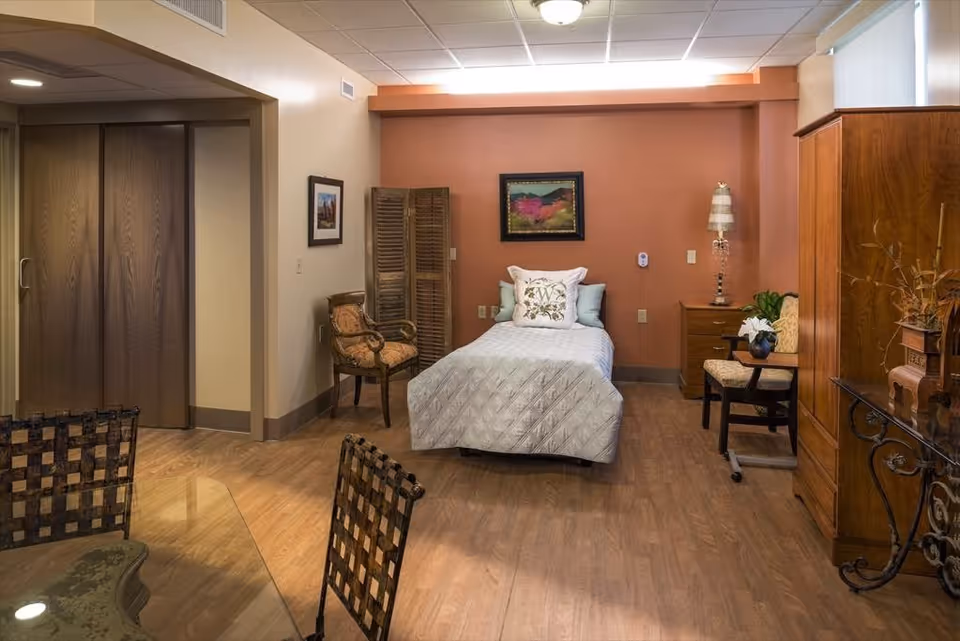 A furnished senior bedroom with a single bed against a coral accent wall, wood furniture, and seating.