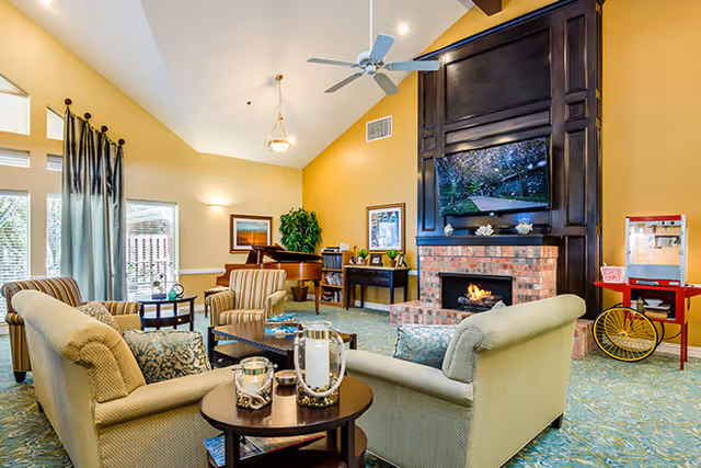 A cozy living room area with beige sofas and armchairs arranged around a coffee table. There is a brick fireplace with a large dark wood mantel and a flat-screen TV mounted above it. A popcorn machine on a red cart is visible to the right. The room has high vaulted ceilings with a ceiling fan and large windows with curtains letting in natural light. There is a piano and framed artwork on the walls, along with a green plant in the corner.