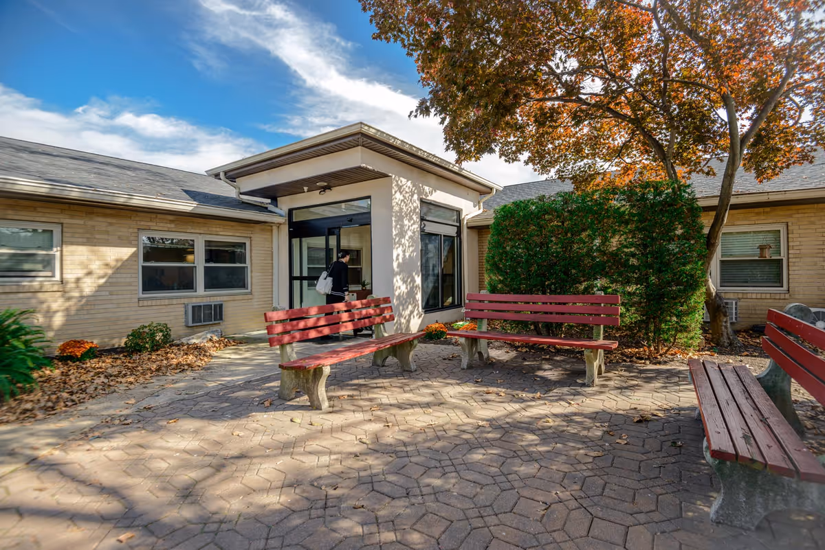 Outdoor courtyard area of a nursing and rehabilitation center with three red wooden benches on a paved surface. There are trees with autumn leaves and bushes around the courtyard. A person is entering the building through a glass door.