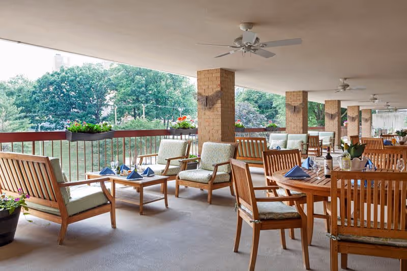 Covered outdoor patio area with wooden chairs, tables set with blue napkins, wine glasses, and potted plants. The patio overlooks a green space with trees in the background.