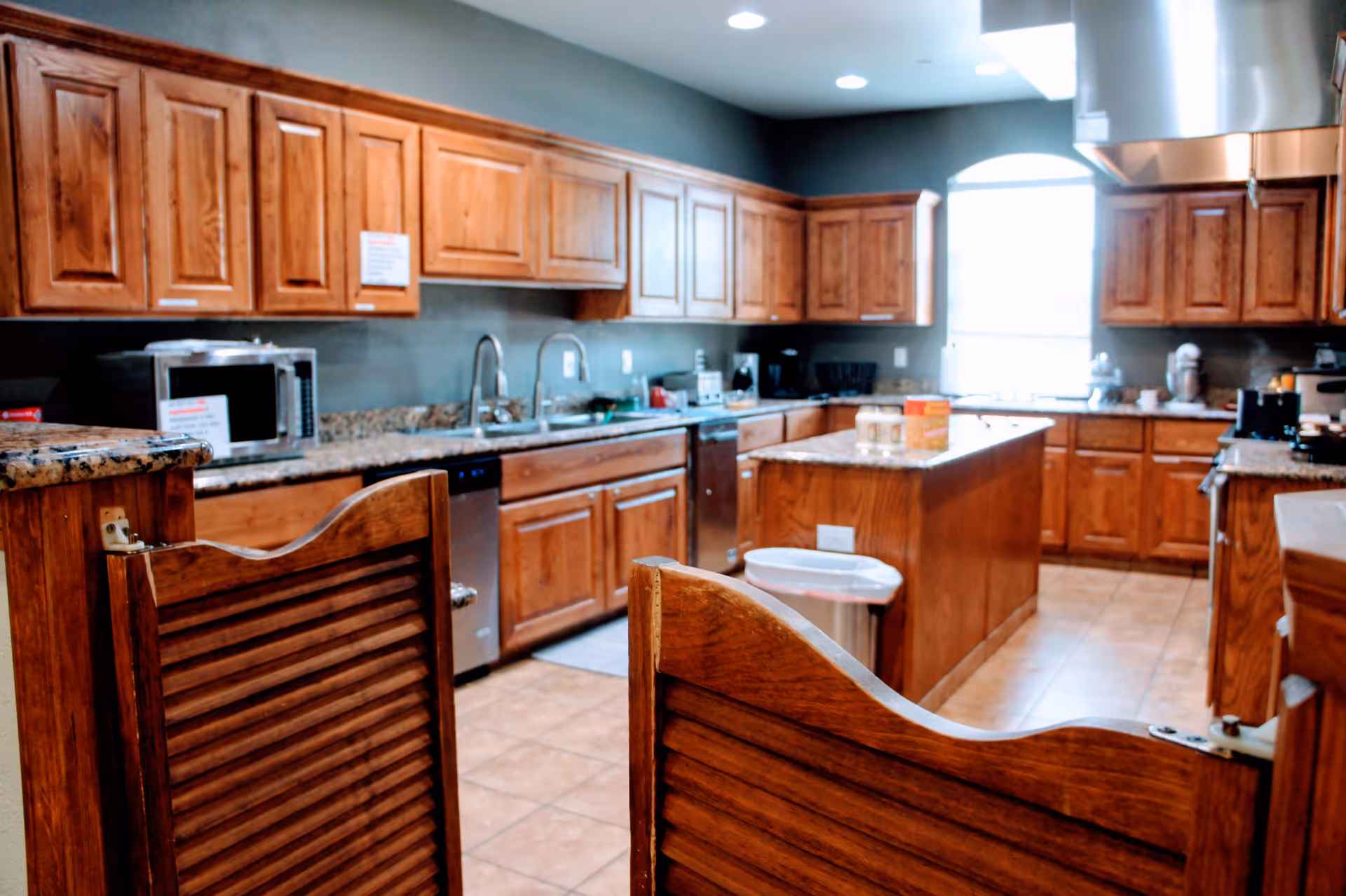 A spacious kitchen with wooden cabinets, granite countertops, a kitchen island, and stainless steel appliances including a microwave and dishwasher. The kitchen has a tiled floor and a window letting in natural light. There is a wooden swinging gate in the foreground.