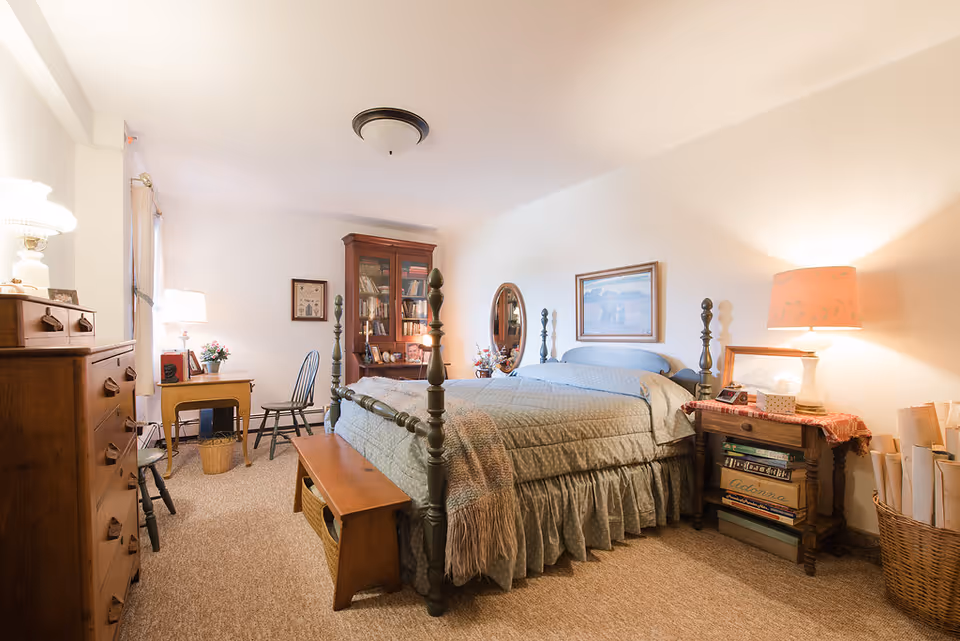 A cozy bedroom with a large bed covered in a light blue quilt and a wooden bench at the foot. The room features a wooden dresser, a small desk with a chair, a tall wooden cabinet filled with books, and a nightstand with a lamp and books. The walls are decorated with framed pictures and a round mirror. The room is warmly lit with lamps and has a carpeted floor.
