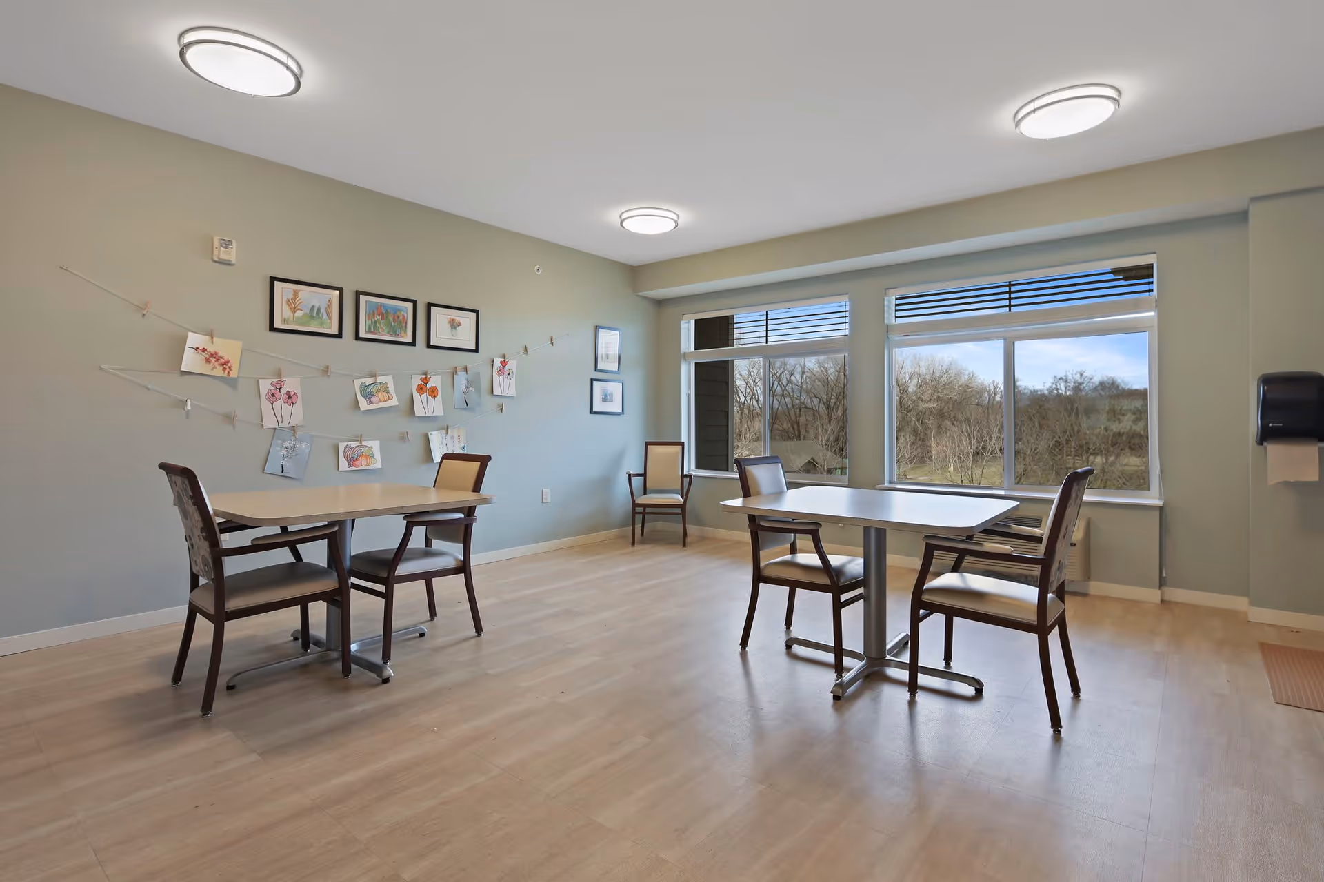A bright room with light wood flooring and pale green walls featuring two tables each surrounded by chairs. The wall has several framed pictures and children's artwork clipped to strings. Large windows show a view of trees outside. Ceiling lights illuminate the space.