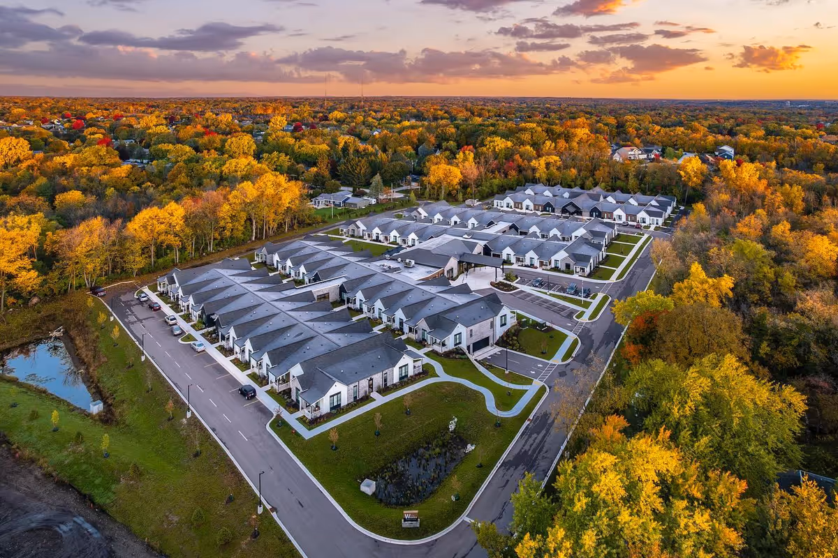 Aerial view of The Woods - A GreatLife Community senior living facility surrounded by trees with autumn foliage. The facility consists of multiple single-story buildings with gray roofs arranged in a neat layout with roads and parking spaces. The sky is partly cloudy with a warm sunset glow.