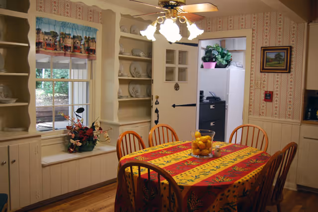 A cozy dining room with a wooden table covered by a colorful red and yellow tablecloth. Six wooden chairs surround the table, which has a glass bowl filled with lemons as a centerpiece. The room features built-in white shelves with decorative plates, a window with a valance, a ceiling fan with lights, and a doorway leading to another room with a filing cabinet and a plant on top.