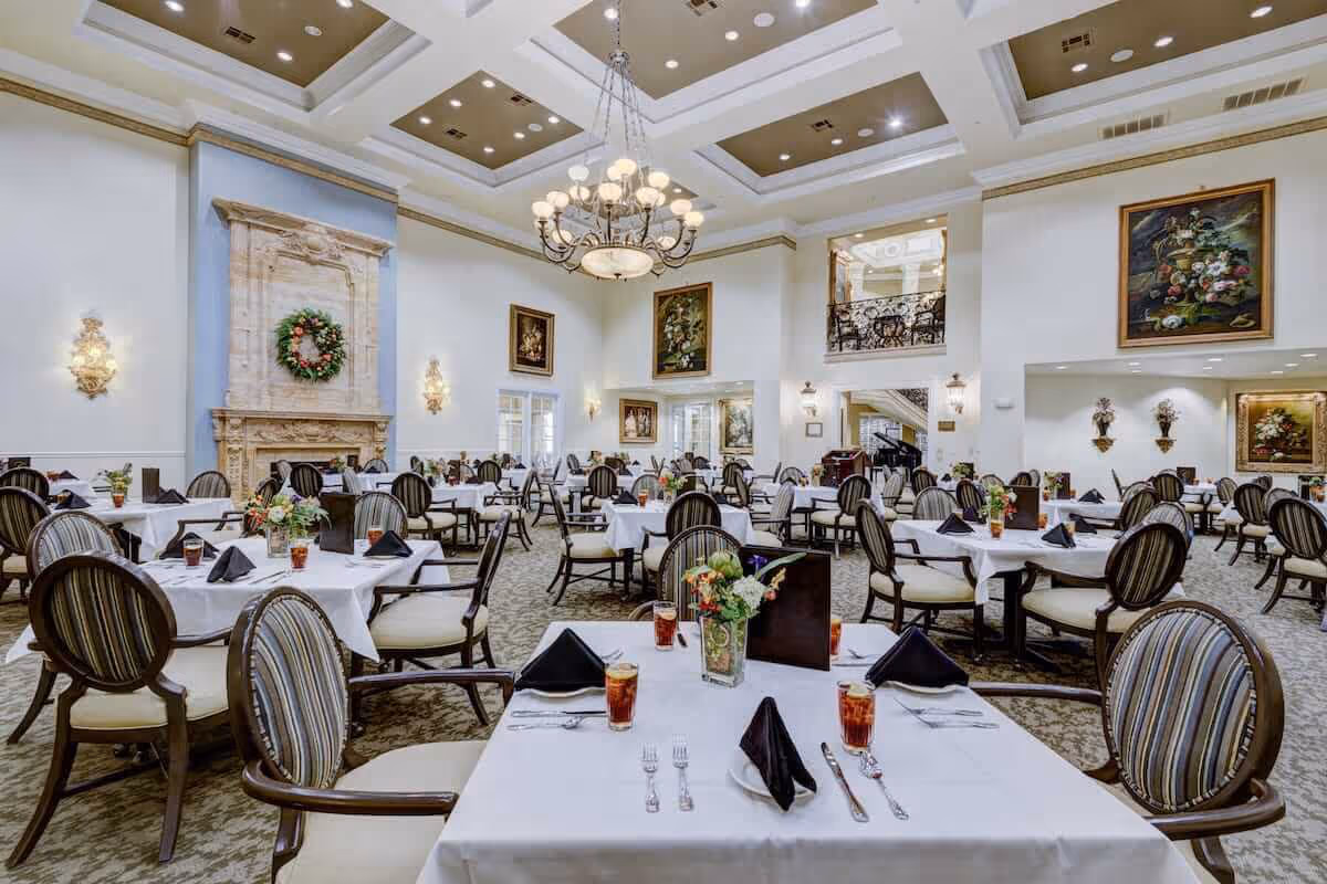 A spacious and elegant dining room with multiple round tables covered in white tablecloths, each set with black napkins, silverware, glasses of iced tea, and floral centerpieces. The room features high ceilings with recessed lighting and a large chandelier, ornate wall sconces, a decorative fireplace with a wreath, and framed paintings on the walls.