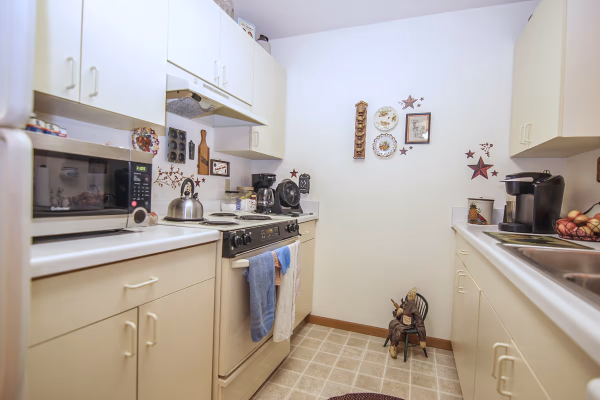 A small kitchen with cream-colored cabinets and white countertops. The kitchen features a microwave, stove with oven, kettle, coffee maker, and a sink. The walls are decorated with star-shaped ornaments, plates, and framed pictures. A small decorative figure is seated on a chair in the corner near the floor.