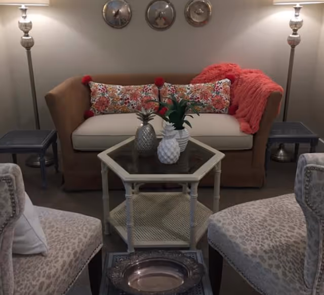 Cozy seating area with a loveseat, patterned throw pillows and a throw, two armchairs, a glass-topped coffee table with decorative pineapple vases, and floor lamps.