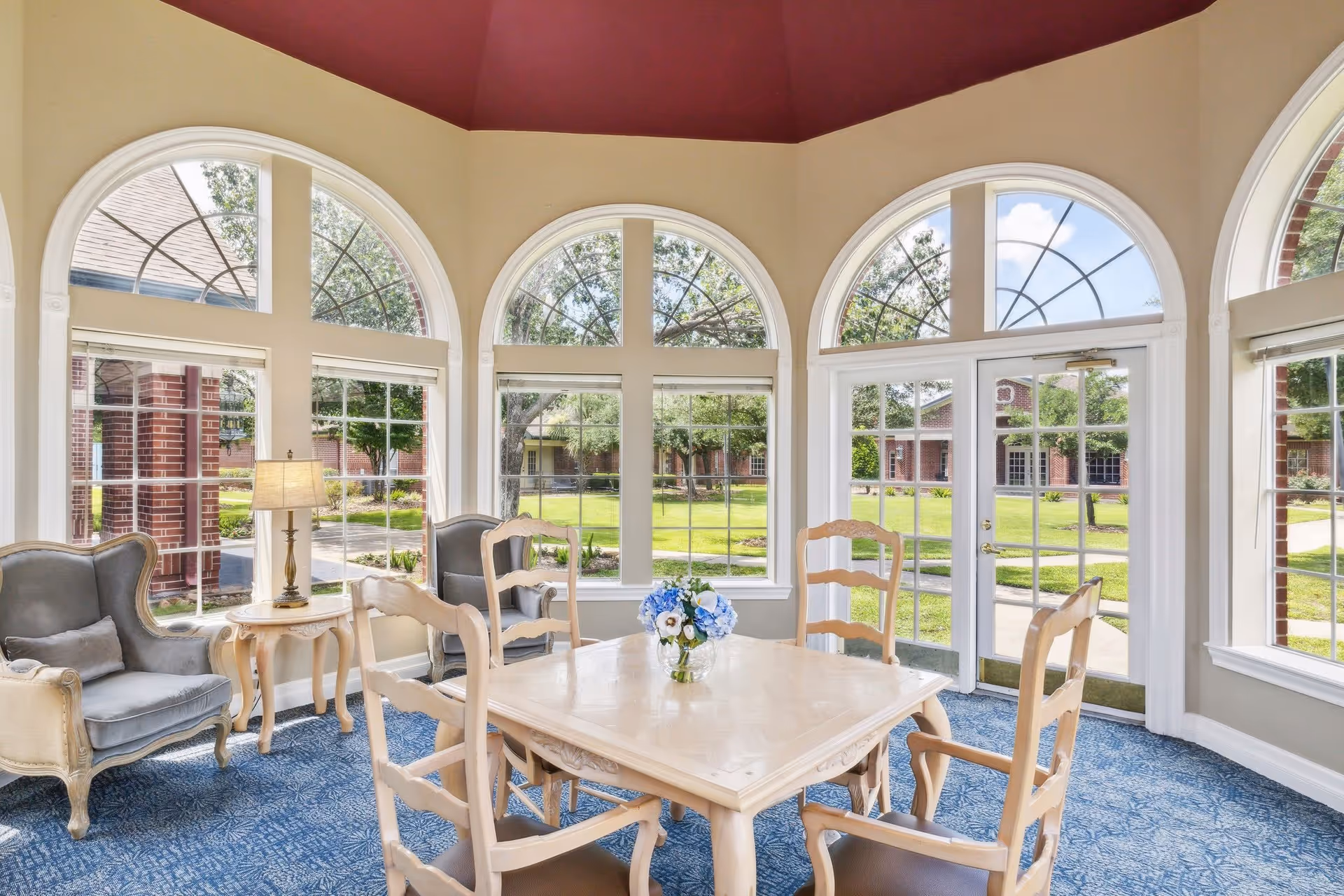 Sunlit common room with a central wooden dining table surrounded by chairs and armchairs in front of large arched windows overlooking a courtyard.