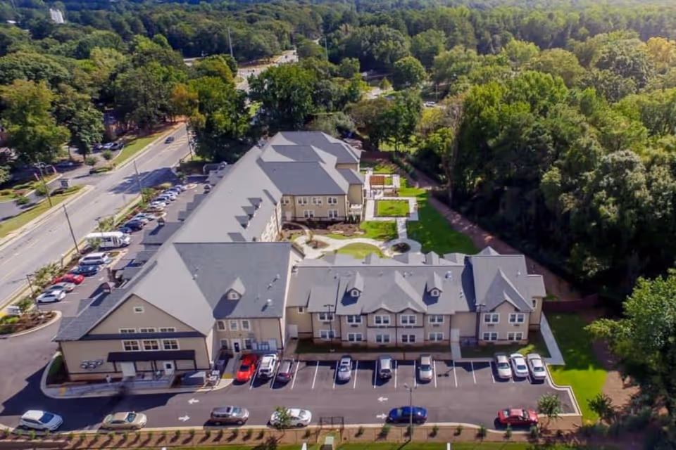 Aerial view of The Phoenix at Tucker senior living facility showing a large two-story building with multiple parking spaces filled with cars, surrounded by trees and greenery under a clear sky.