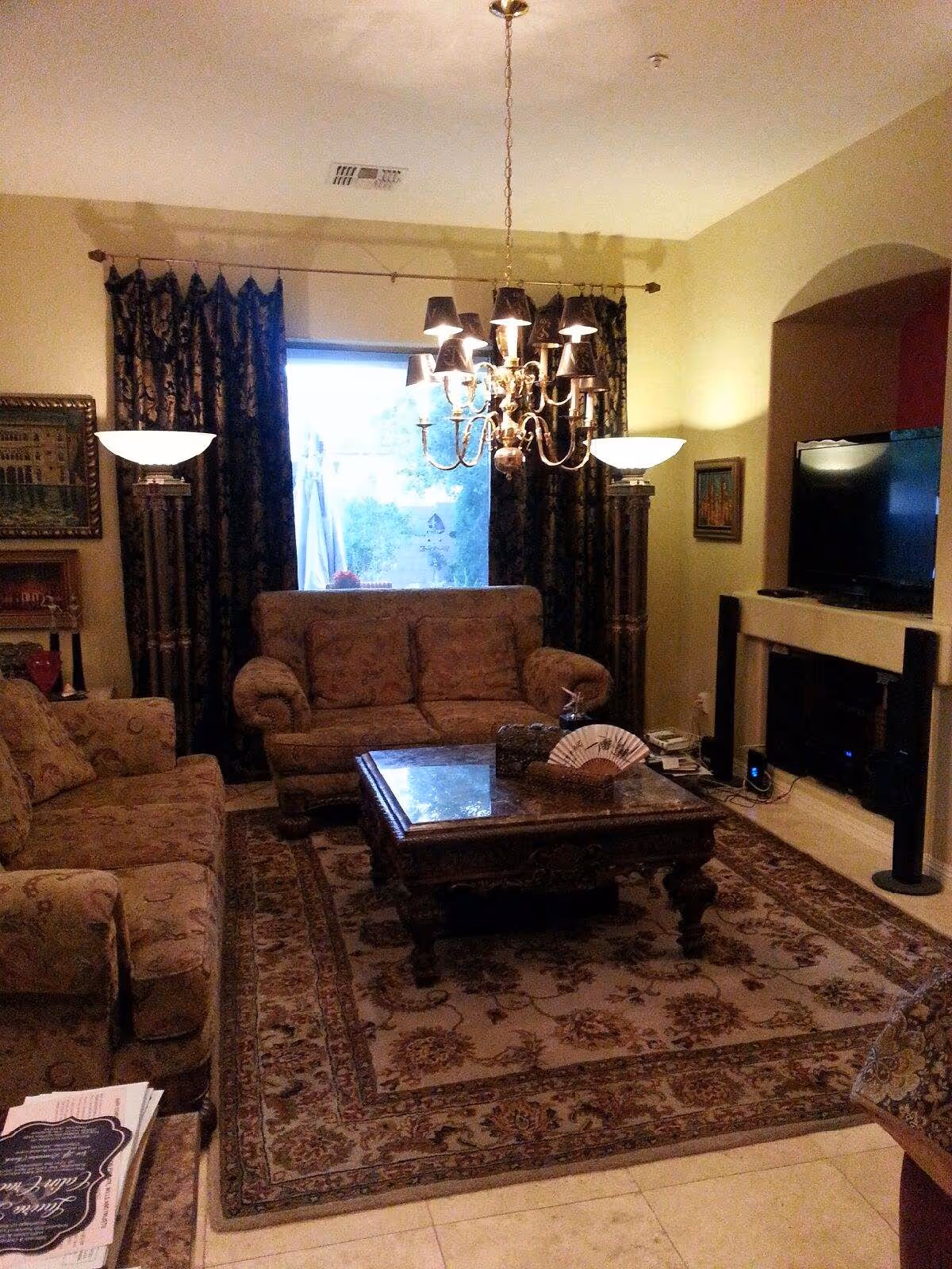 Cozy living room with upholstered sofas and armchairs around an ornate coffee table on a patterned rug, lit by a chandelier with a TV and fireplace in the background.