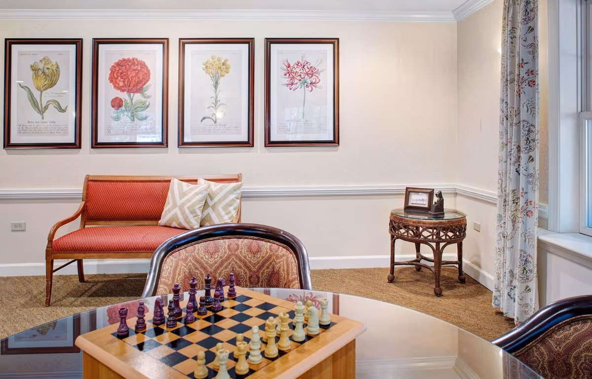 A cozy sitting area in a senior living facility with a wooden chessboard on a glass table in the foreground, two upholstered chairs, a red cushioned bench with two patterned pillows against the wall, four framed botanical prints hanging above the bench, a small round wicker side table with a framed photo and decorative item, and a window with floral curtains on the right.
