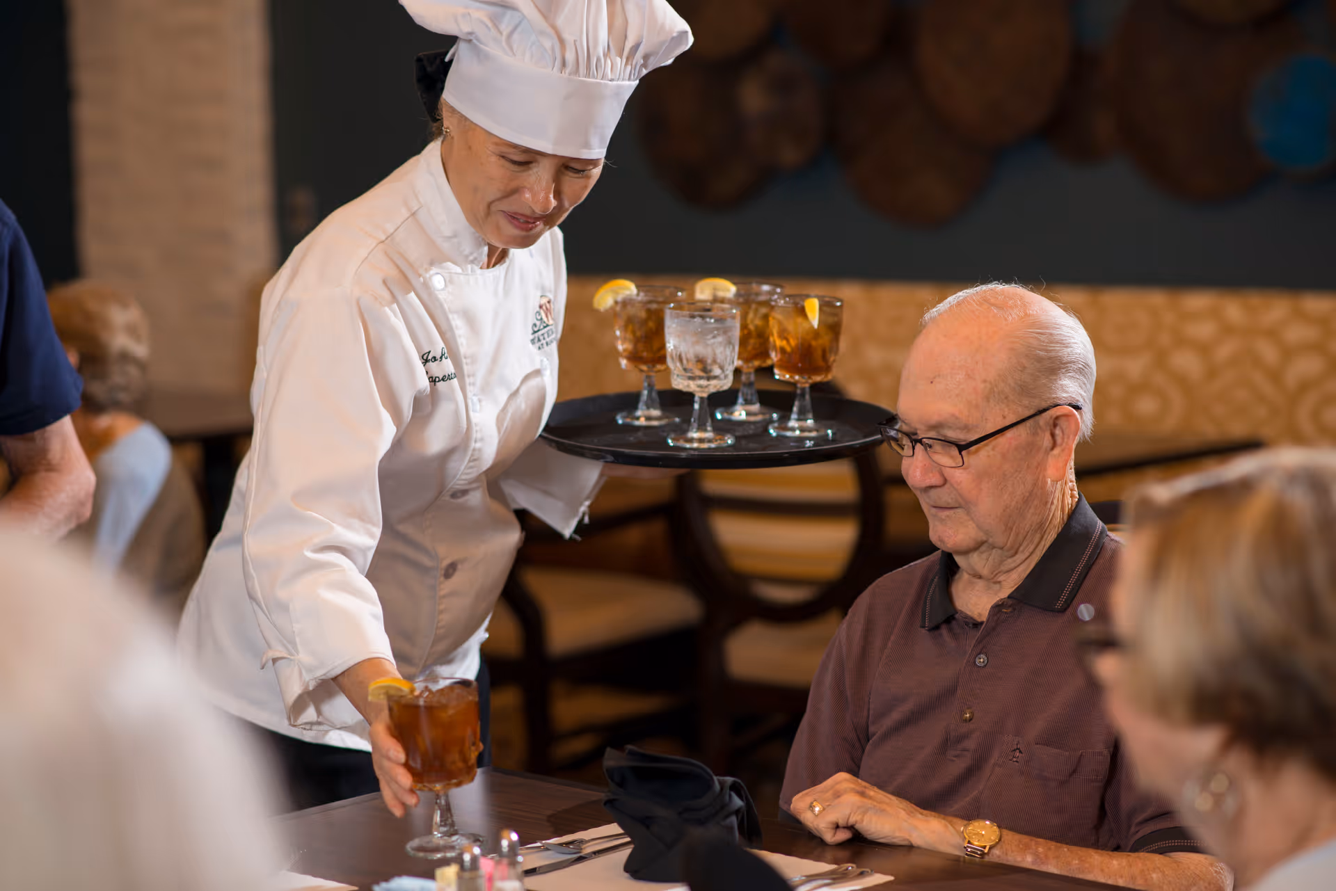 A chef in a white uniform and hat serving iced tea with lemon to an elderly man seated at a dining table in a restaurant setting, with other elderly people visible in the background.