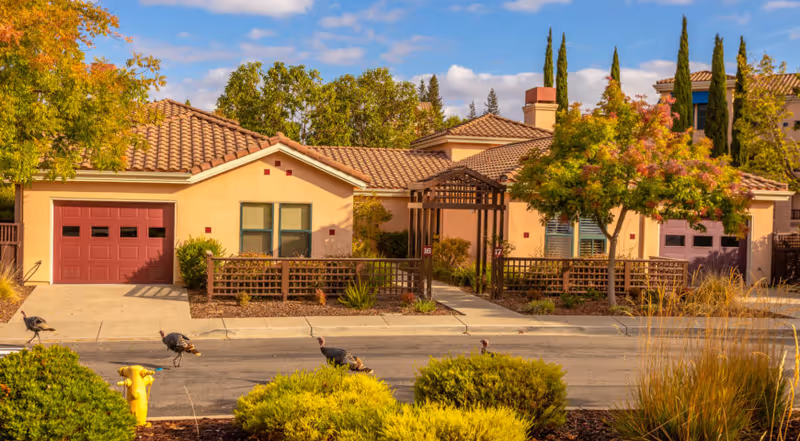 Exterior view of a residential senior living facility with beige stucco walls and red tile roofs. The scene shows two attached units with red garage doors, a wooden pergola entrance, and landscaped greenery including bushes, trees with autumn foliage, and ornamental grasses. Several wild turkeys are walking on the street in front of the units under a partly cloudy blue sky.