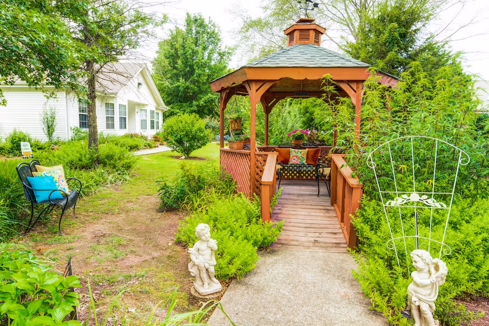 A peaceful garden area at The Renaissance of Florence featuring a wooden gazebo with seating inside, surrounded by lush greenery and decorative statues. There is a black metal bench with colorful cushions on the left side and a white building with multiple windows in the background.