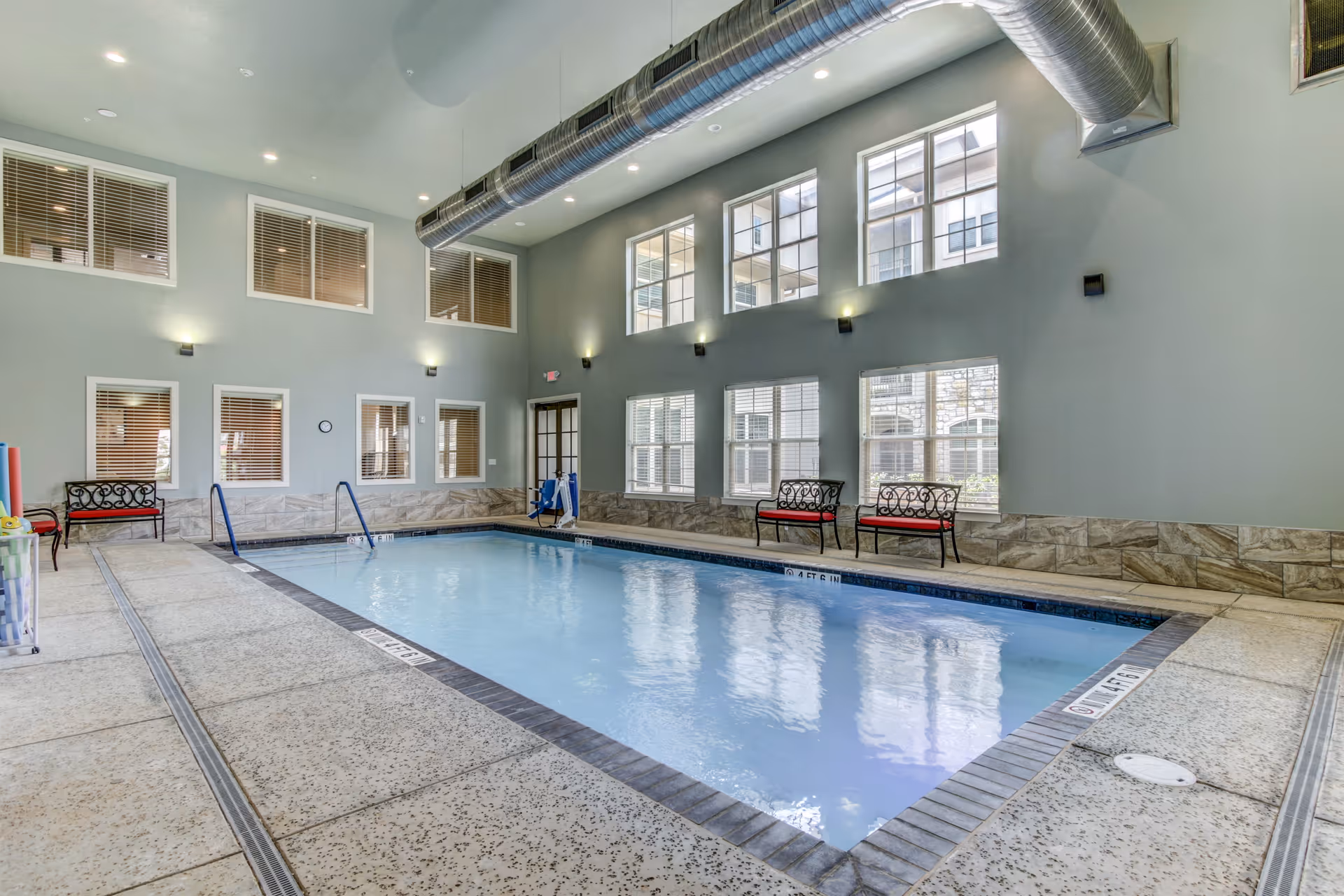 Indoor swimming pool with clear water surrounded by a tiled deck. The room has high ceilings with exposed ductwork and multiple large windows letting in natural light. There are benches with red cushions along the walls and pool safety equipment visible near the pool.