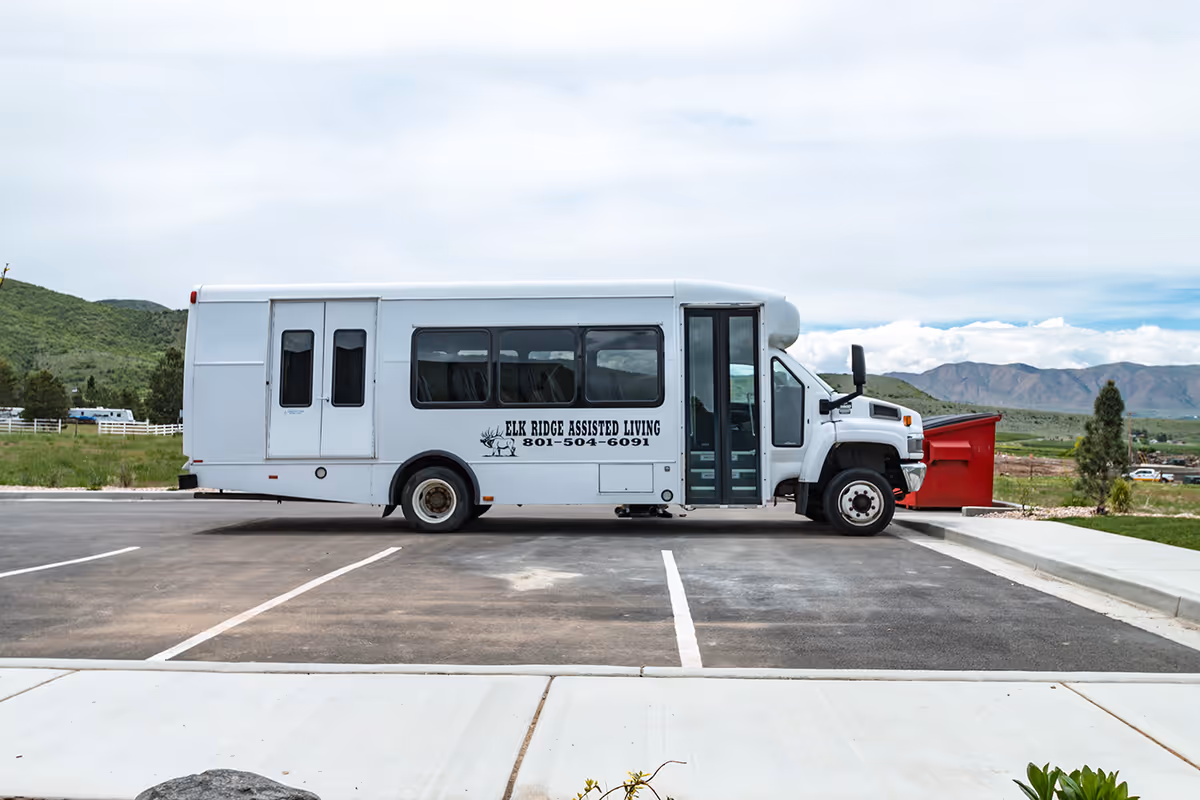 A white Elk Ridge Assisted Living shuttle bus parked in an empty parking lot with mountains in the background.