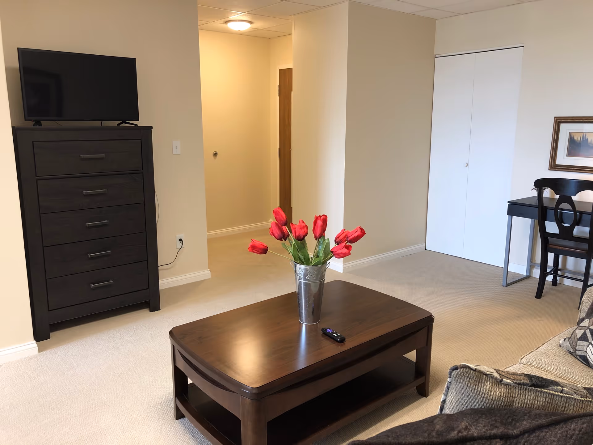 A cozy living room area in The Whitcomb Senior Living Tower featuring a dark wooden coffee table with a metal vase holding red tulips, a TV on a dark wooden dresser, a black desk with a chair, a framed picture on the wall, and a beige carpeted floor.