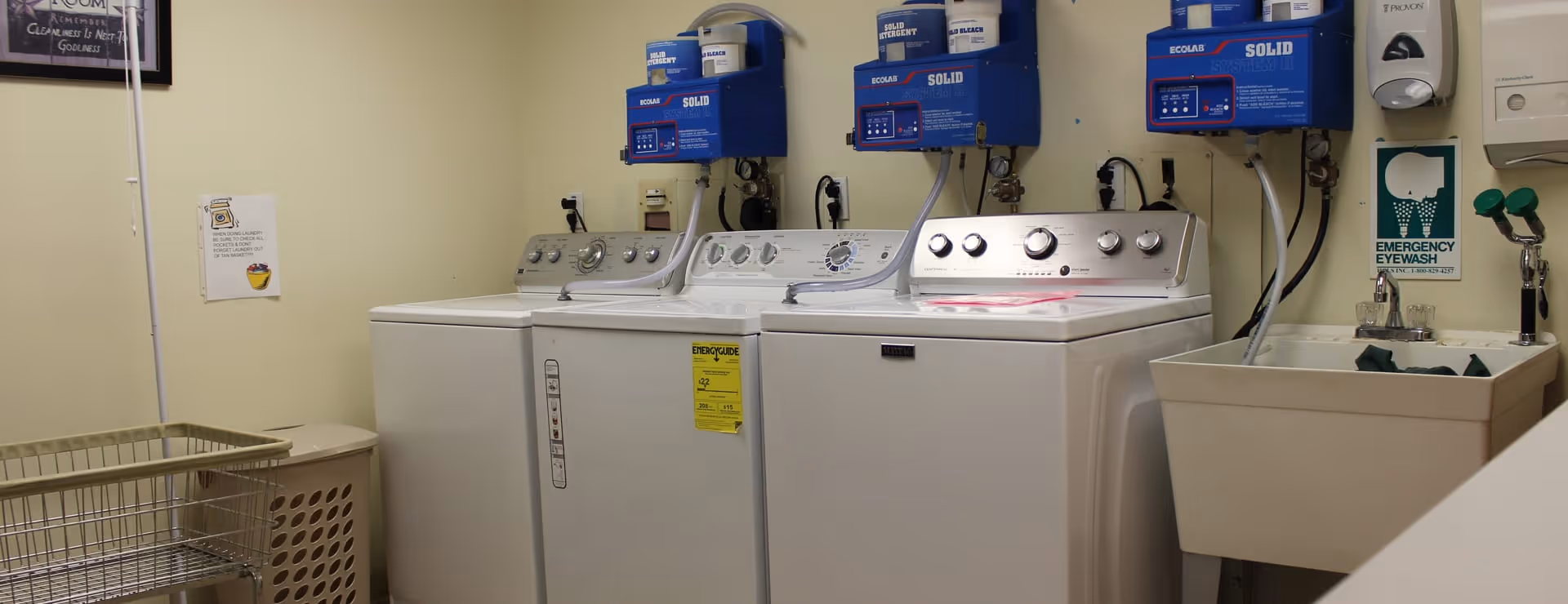 Laundry room with three white washing machines and dryers, a laundry basket, a beige hamper, an emergency eyewash station, and cleaning supply dispensers mounted on the wall.