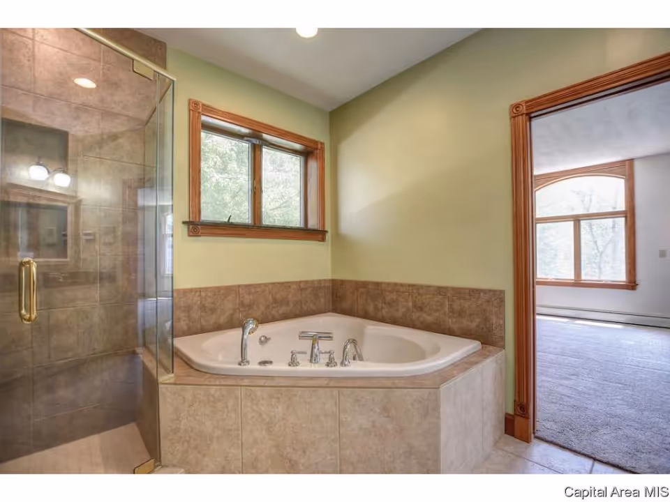 Bathroom with a corner bathtub featuring multiple faucets and a tiled surround. Next to the bathtub is a glass-enclosed shower with a brass handle. The walls are painted light green and there is a window with wooden trim above the bathtub. An open doorway with wooden trim leads to a carpeted room with large windows.