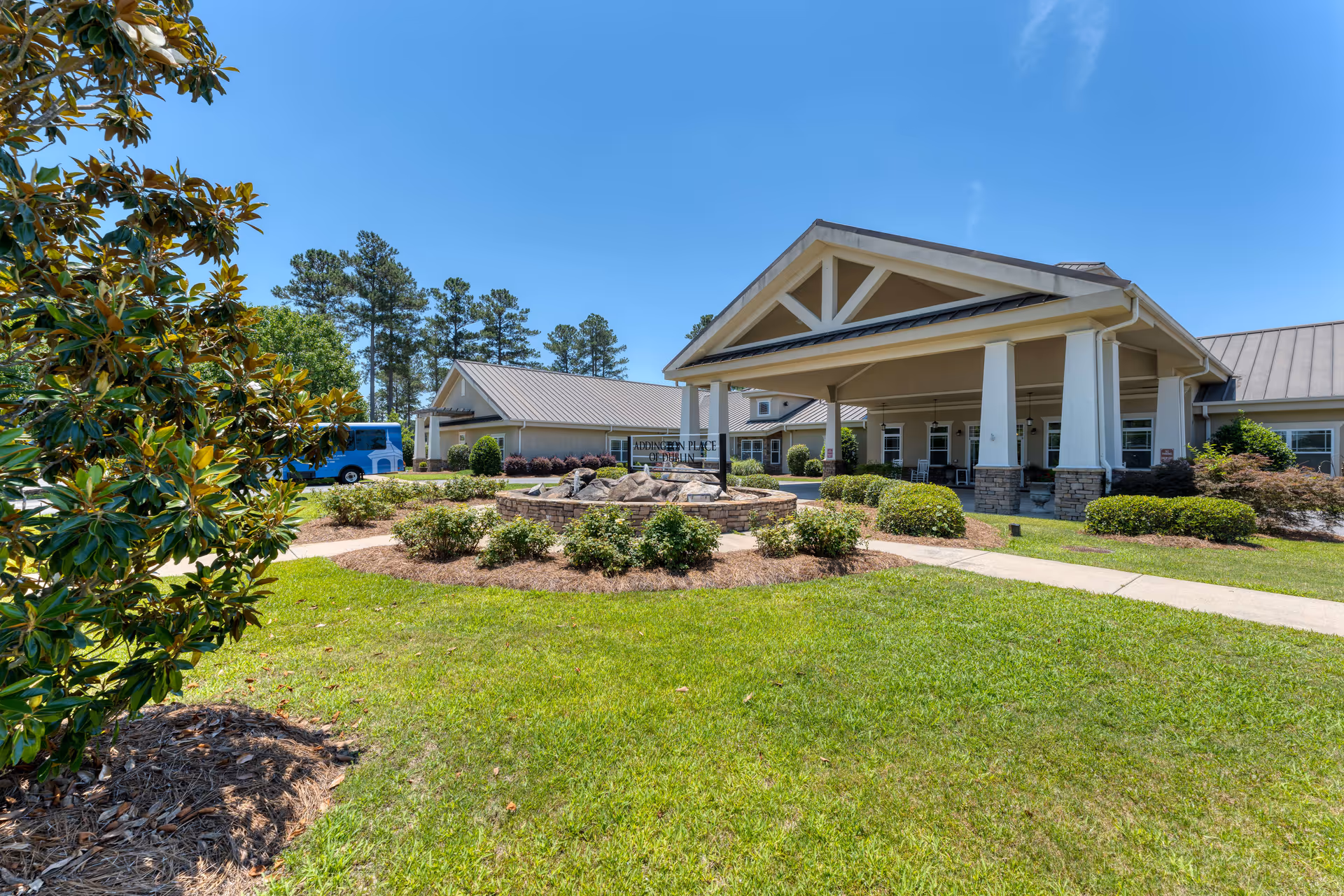 Exterior view of Addington Place of Dublin senior living facility on a sunny day, showing a large covered entrance with white pillars, manicured bushes, green lawn, and a clear blue sky. A blue shuttle bus is parked near the building.