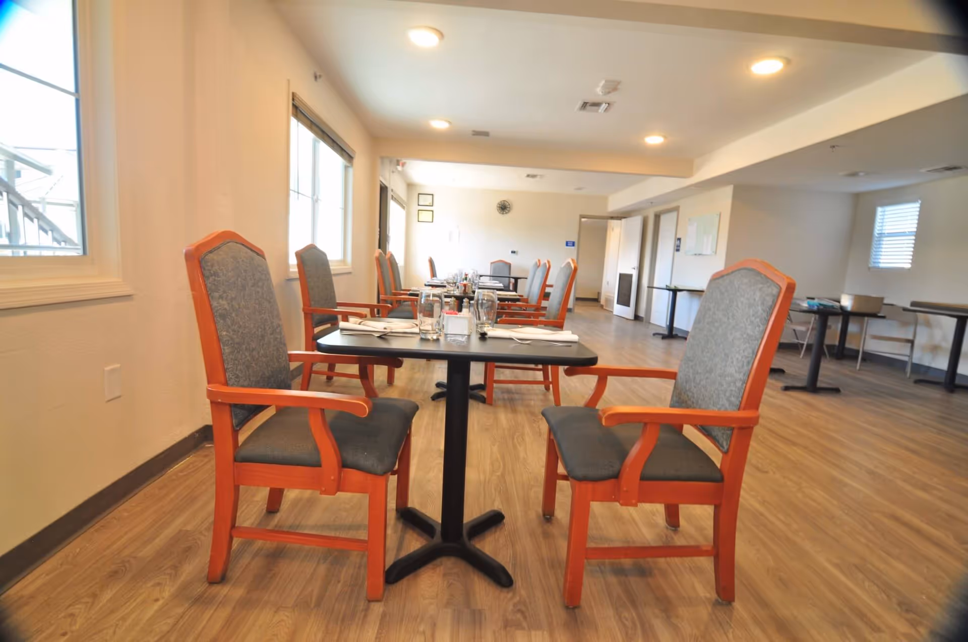 A dining room with multiple tables and chairs arranged in rows. Each table has two chairs with wooden frames and cushioned seats and backs. The tables are set with glasses, napkins, and condiments. The room has large windows on one side allowing natural light to enter, and the floor is covered with wood-patterned flooring. The walls are light-colored, and there are ceiling lights providing additional illumination.