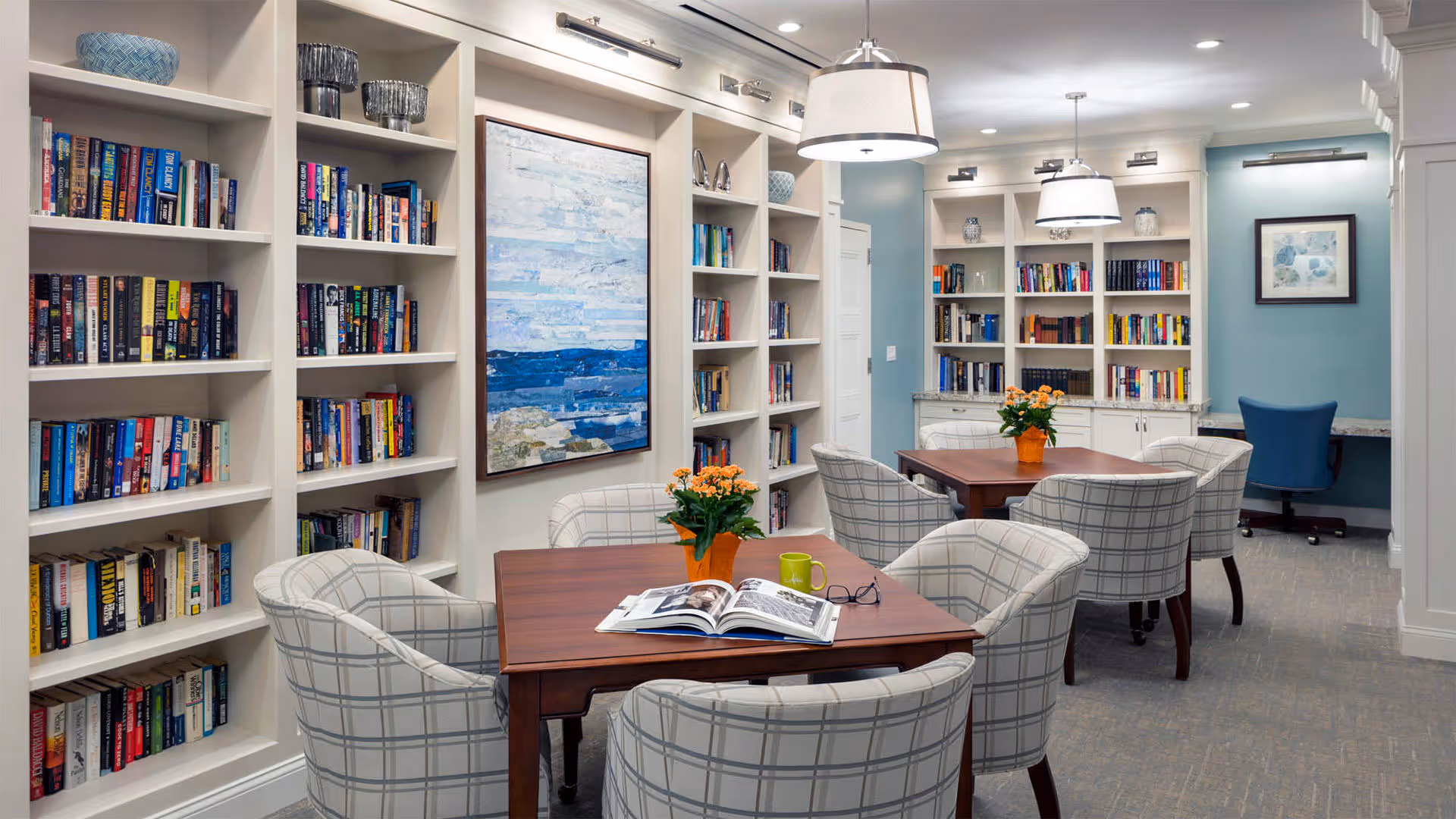 A cozy reading room with built-in white bookshelves filled with books, two wooden tables each surrounded by four cushioned chairs with a plaid pattern. There are orange flower pots on the tables, a large painting of a seascape on the wall, and a blue accent wall with additional bookshelves and a desk with a blue chair. The room is well-lit with ceiling lights and wall-mounted picture lights.