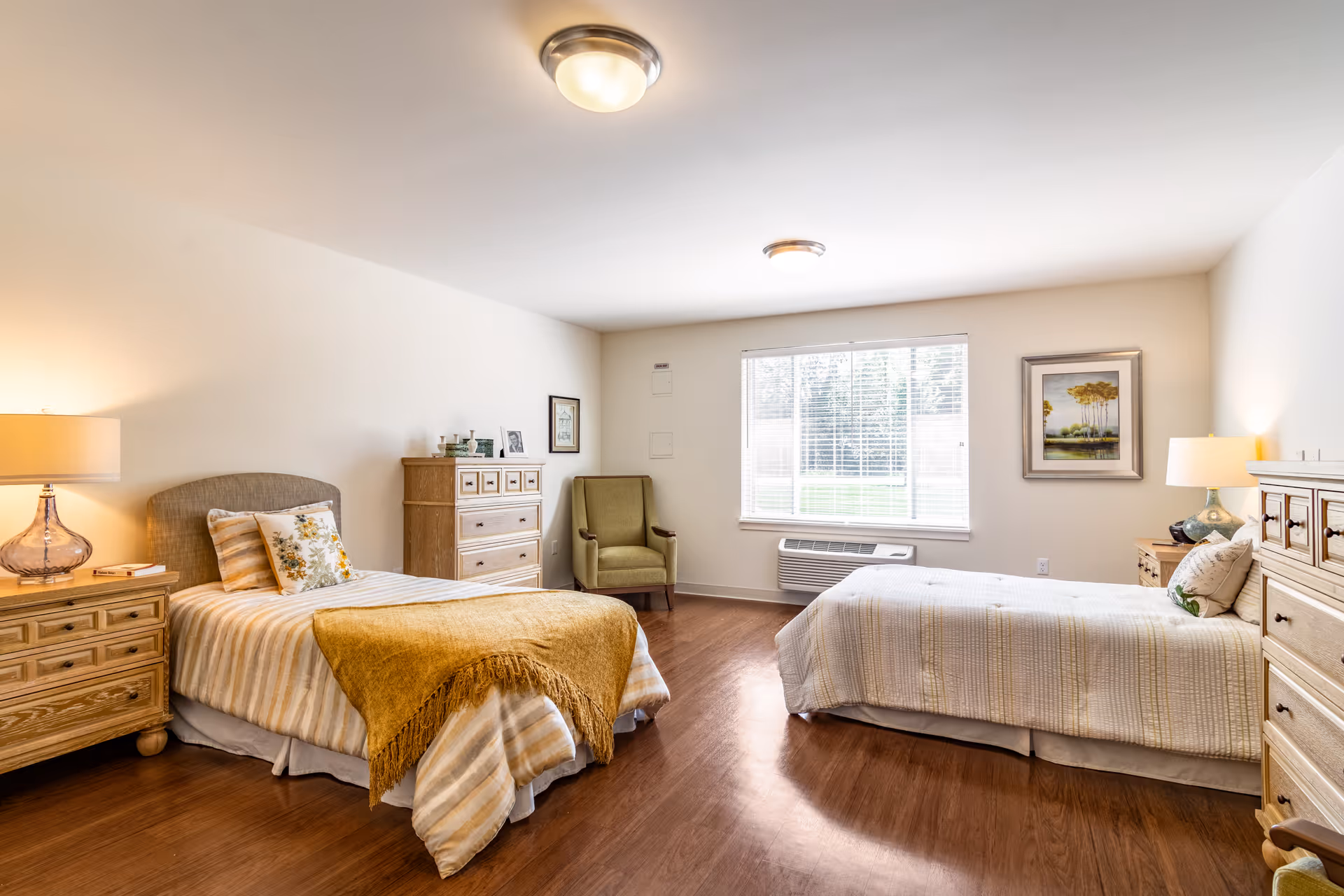 A bright and spacious bedroom with two single beds, each with a bedside table and lamp. The bed on the left has a yellow throw blanket and floral pillow, while the bed on the right has a white quilt and decorative pillow. Between the beds is a large window with blinds, and a green armchair is positioned in the corner. There are two wooden dressers, one on each side of the room, and framed artwork on the walls. The floor is wooden, and the room has a clean, cozy atmosphere.