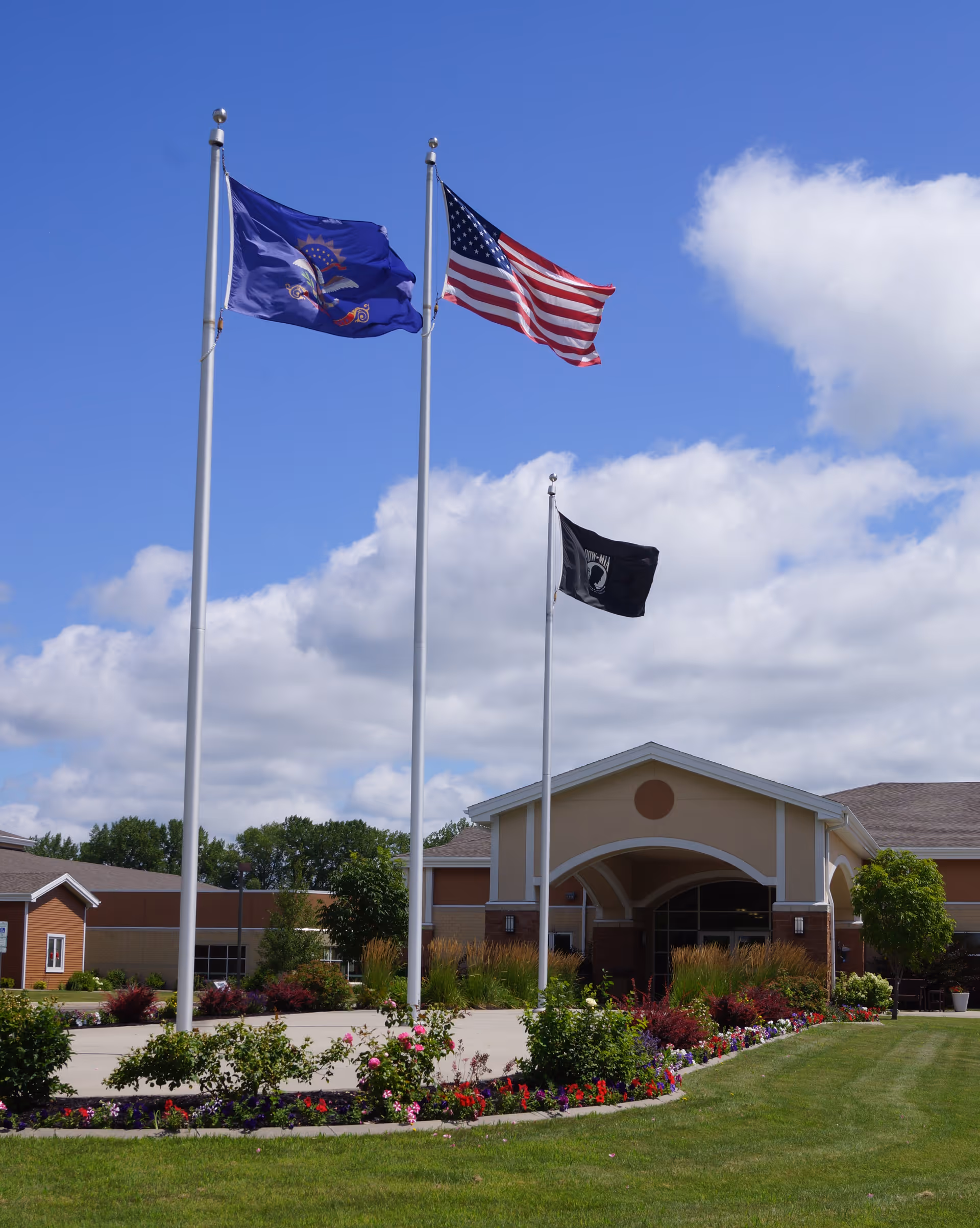 Three flagpoles with flags flying in front of a single-story building with an arched entrance. The flags include the North Dakota state flag, the United States flag, and a black POW/MIA flag. The area around the flagpoles is landscaped with green grass, bushes, and colorful flowers under a partly cloudy blue sky.