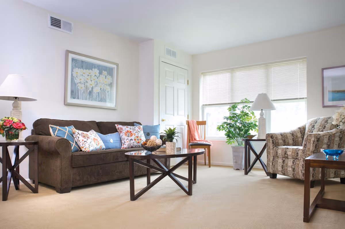 A bright and cozy living room with a brown sofa adorned with colorful patterned pillows, a patterned armchair, a wooden coffee table with decorative items, side tables with lamps and flowers, a wooden chair with a pink throw, and a large window with blinds letting in natural light. The walls are light-colored with framed artwork and a potted plant near the window.