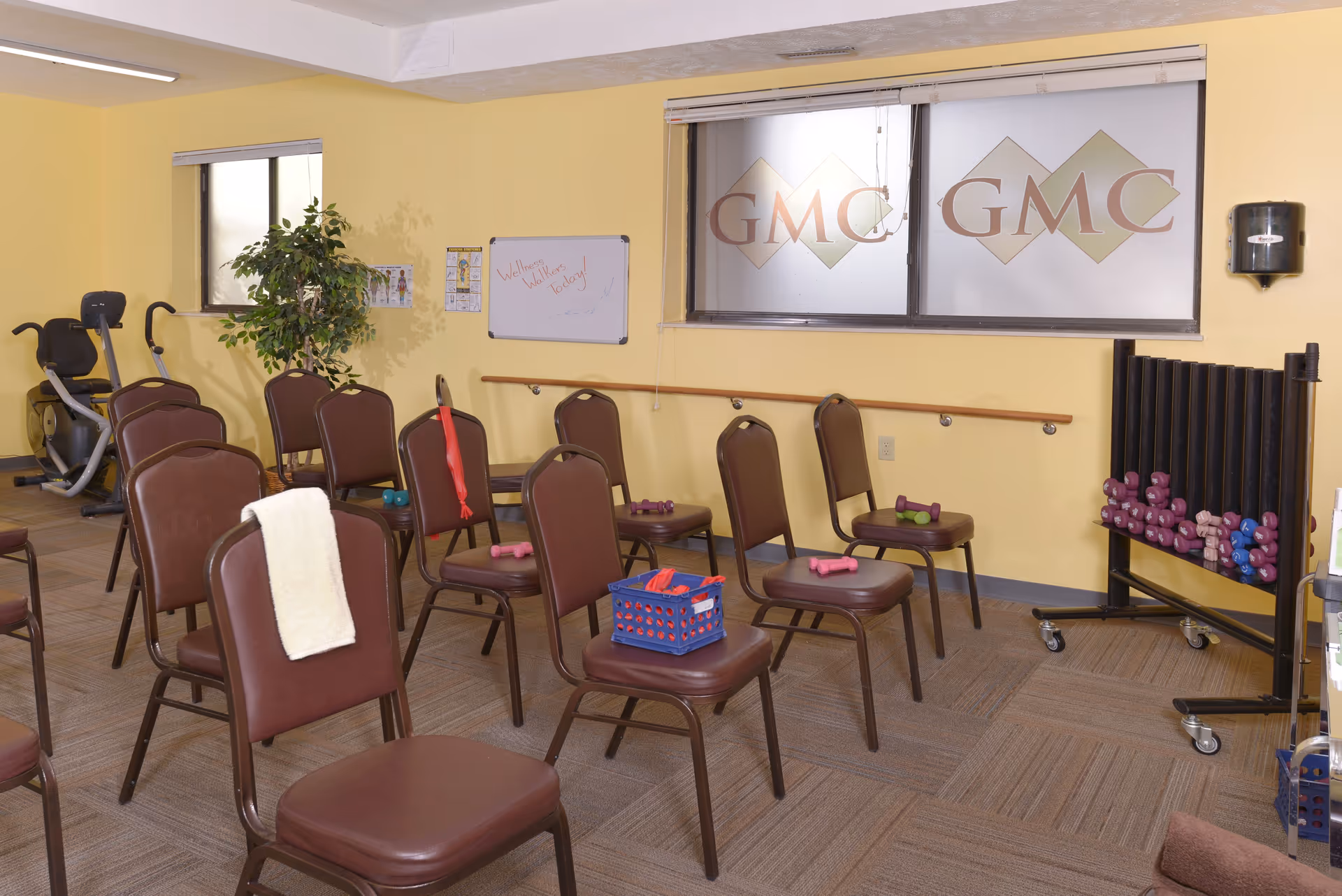 A small exercise room with several brown chairs arranged in rows, each chair holding small dumbbells or resistance bands. There is a stationary exercise bike in the back left corner, a potted plant, a whiteboard on the wall with the message 'Wellness Walkers Today!', and a window with frosted glass displaying the letters GMC. A rack with more dumbbells is on the right side of the room.
