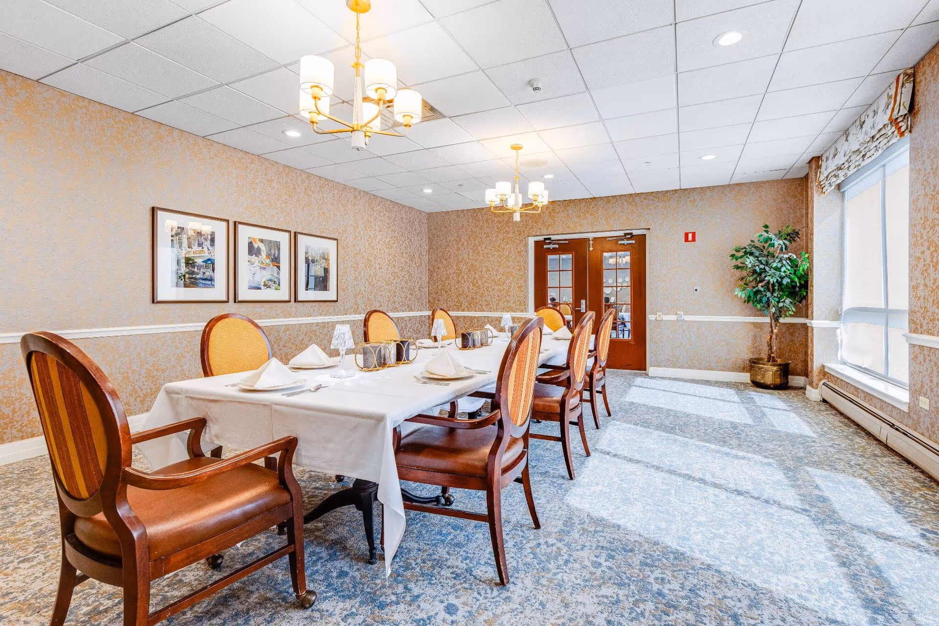 A bright dining room with a long set table, upholstered wooden chairs, chandeliers, and a large window letting in sunlight.