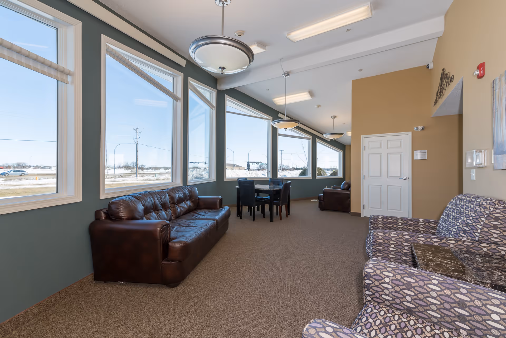 Bright common lounge with a leather sofa, patterned armchairs, and a table set by large windows.