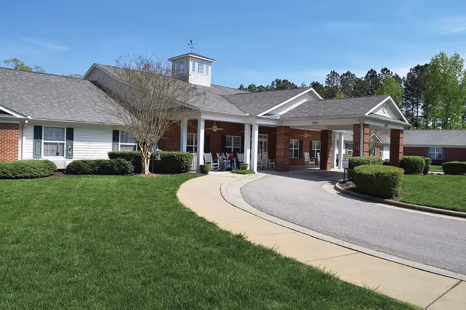 Exterior view of Spring Arbor of Apex facility showing a single-story building with a covered entrance supported by brick columns, surrounded by green grass, bushes, and trees under a clear blue sky.