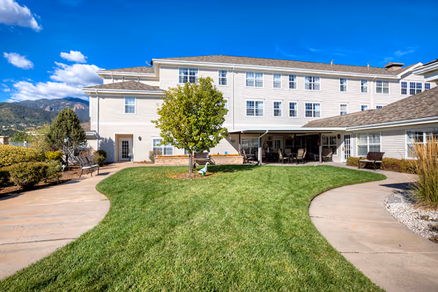 Outdoor view of Bear Creek Assisted & Senior Living facility showing a well-maintained green lawn with a tree in the center, surrounded by paved walkways and benches. The building is a multi-story structure with white siding and multiple windows under a bright blue sky with some clouds.