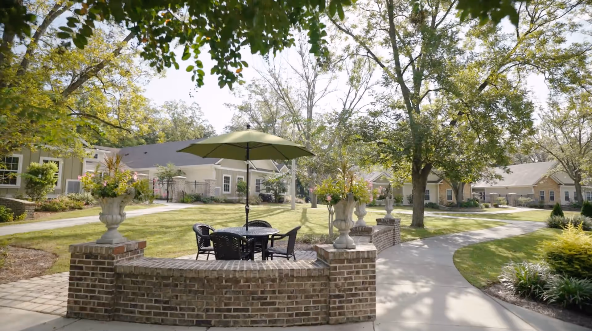 Outdoor seating area with a round table, four black chairs, and a green umbrella surrounded by a low brick wall with flower pots. The area is set in a garden with trees, grass, and paved walkways leading to single-story buildings in the background.