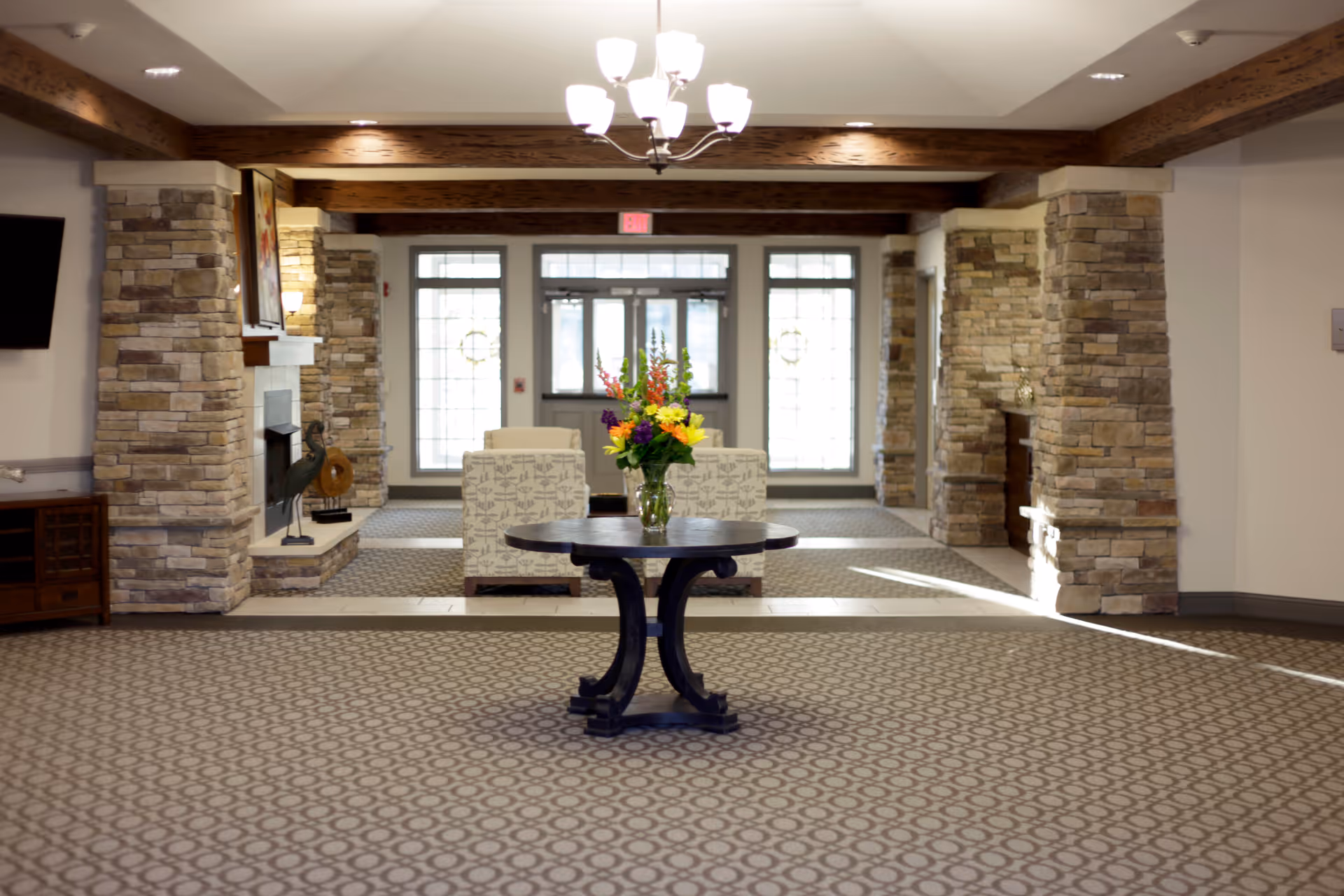 A spacious and well-lit interior common area with patterned carpet flooring, stone pillars, and wooden ceiling beams. In the center, there is a round wooden table with a vase of colorful flowers. In the background, there are two upholstered armchairs facing a set of glass doors and windows allowing natural light to enter.