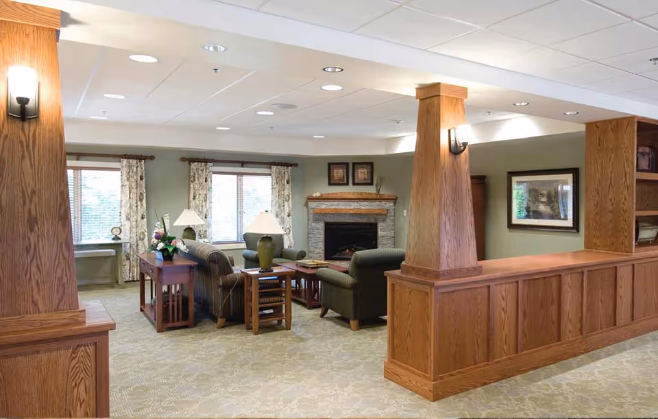 A cozy living room area in a retirement community featuring wooden pillars and paneling, a stone fireplace, two armchairs, a sofa, side tables with lamps, floral curtains on windows, and framed artwork on the walls.