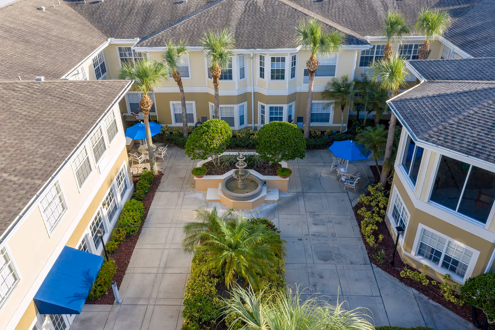 Aerial view of a courtyard at Aston Gardens At The Courtyards featuring a central water fountain, neatly trimmed bushes, palm trees, outdoor seating with tables and blue umbrellas, surrounded by a two-story building with multiple windows.