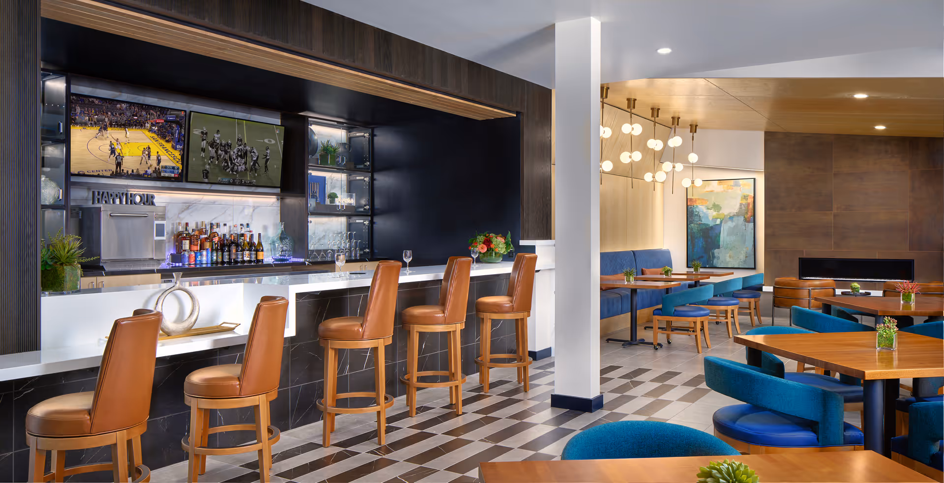 Modern senior living facility lounge area featuring a bar with four brown leather bar stools, two televisions mounted above the bar showing sports games, and a seating area with blue cushioned chairs and wooden tables. The room has contemporary lighting fixtures, decorative plants, and a fireplace in the background.