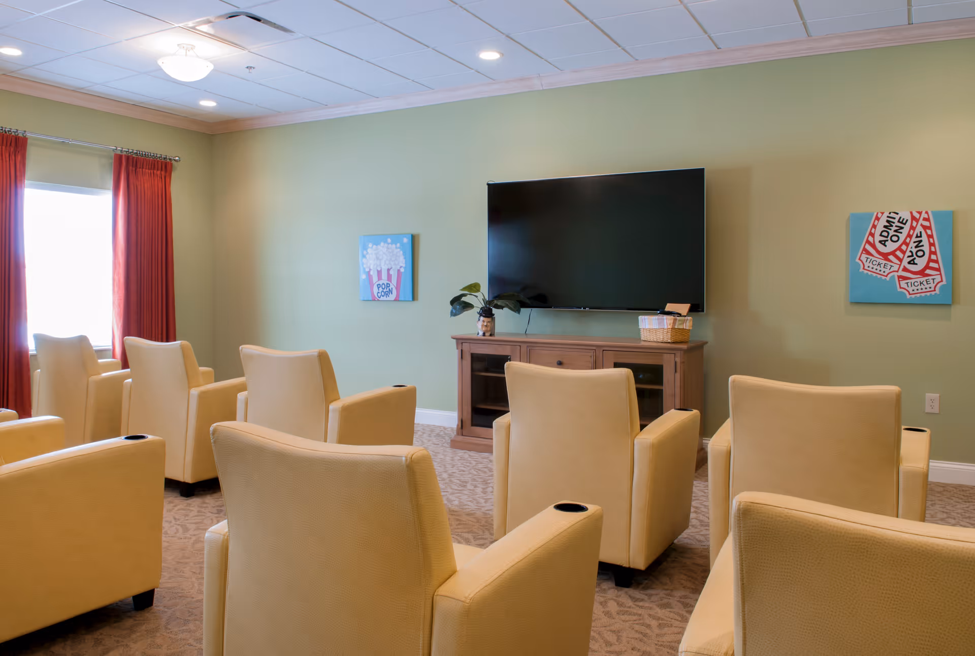 A small theater-style room with six beige armchairs arranged in two rows facing a large flat-screen TV mounted on a light green wall. The room has a beige carpet, red curtains on a window to the left, and two colorful paintings on the wall, one depicting popcorn and the other showing admission tickets. A wooden cabinet sits below the TV with a small plant and a basket on top.