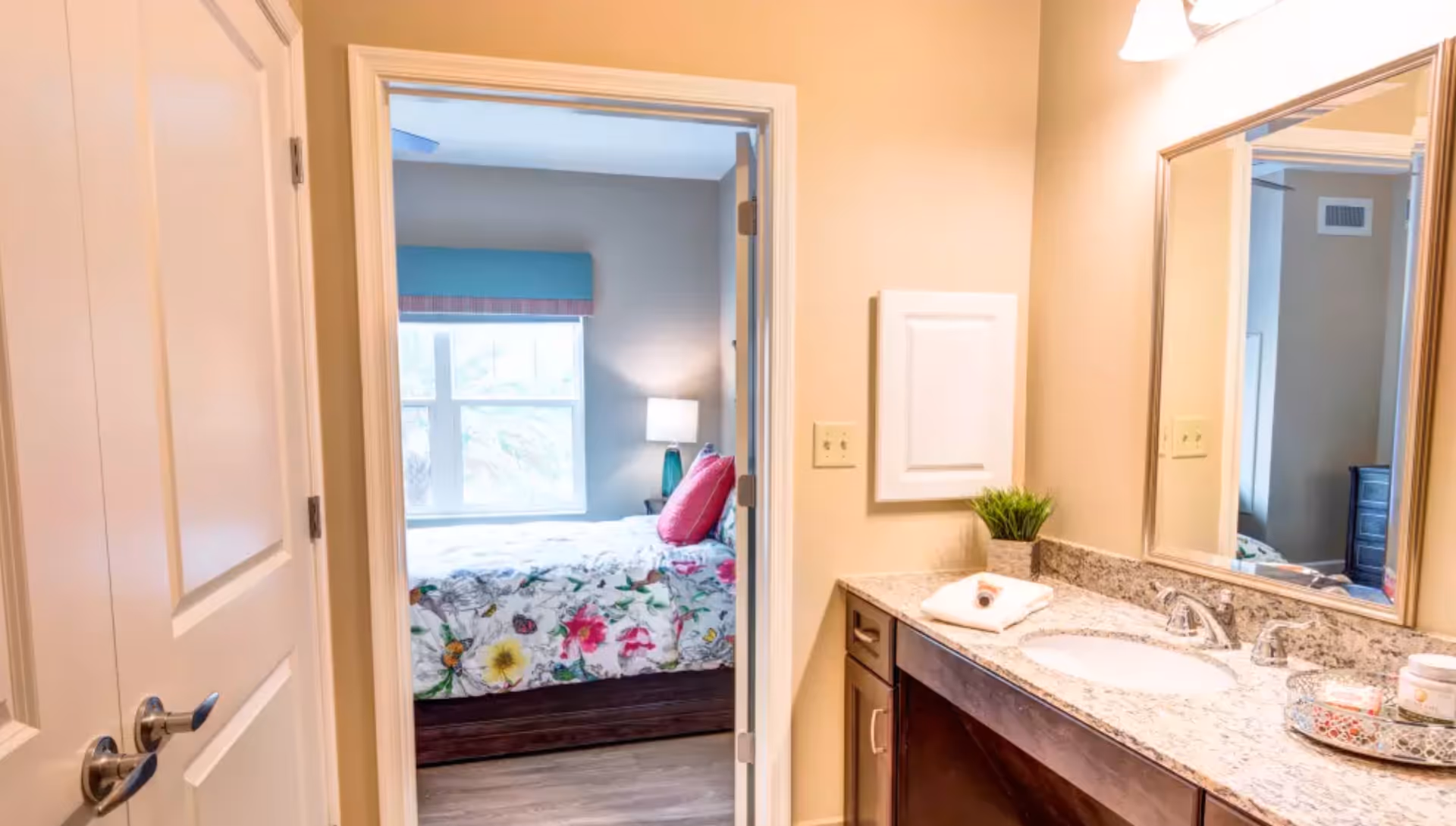 Bathroom vanity and mirror with a doorway leading to a bedroom featuring a floral bedspread and window.