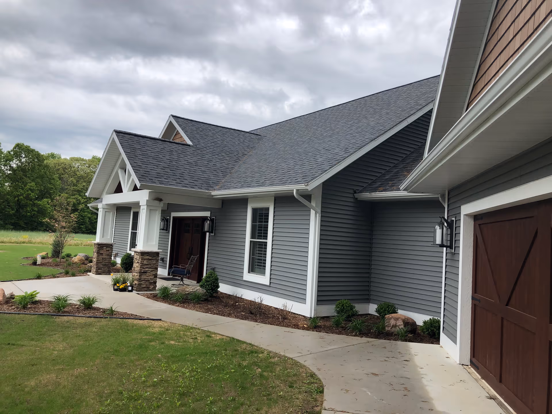 Exterior view of a single-story building with gray siding, white trim, and a dark shingled roof. The entrance features a covered porch with stone pillars and outdoor seating. There is a concrete walkway leading to the entrance, surrounded by landscaped grass and small shrubs. The sky is overcast.