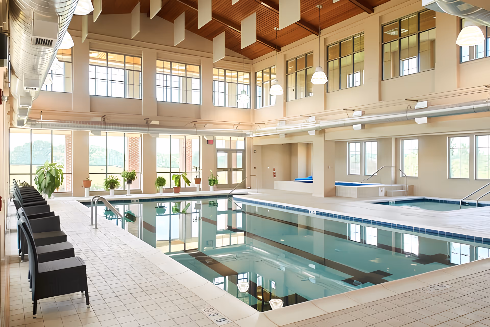 Spacious, sunlit indoor pool area with lounge chairs, potted plants, large windows, and an adjacent hot tub.