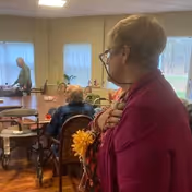 An elderly woman wearing glasses and a magenta shawl stands in the foreground of a room with several elderly people seated at tables. The room has large windows with sheer curtains, and there are chairs and tables arranged for social or dining activities.