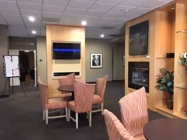 Common lounge with round tables, patterned upholstered chairs, a wall-mounted TV and a fireplace.