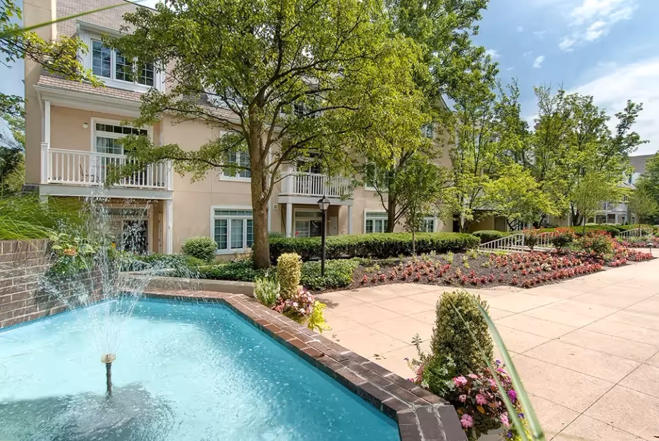 Outdoor view of a senior living facility with a small water fountain in the foreground, surrounded by a brick border. There are well-maintained flower beds, shrubs, and trees along a paved walkway. The building in the background has balconies and large windows, with a clear blue sky above.