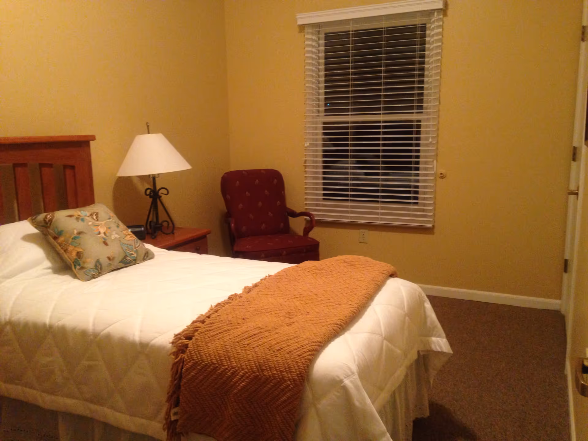 A cozy bedroom with a single bed covered in a white quilt and an orange throw blanket. There is a decorative pillow on the bed. Next to the bed is a wooden nightstand with a lamp and an alarm clock. A red upholstered armchair is placed near a window with white blinds. The walls are painted beige and the floor is carpeted.